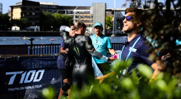 A triathlete in a wetsuit is helped by staff during a triathlong transition. The T100 Triathlon World Tour banner is visible.