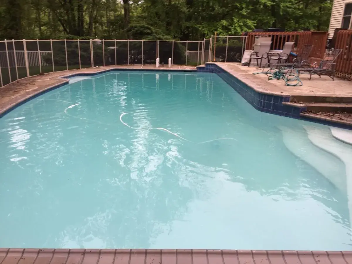 A backyard pool with cloudy, turquoise water, a white hose, and a safety fence. You can swim in this cloudy pool, but it might not be ideal.