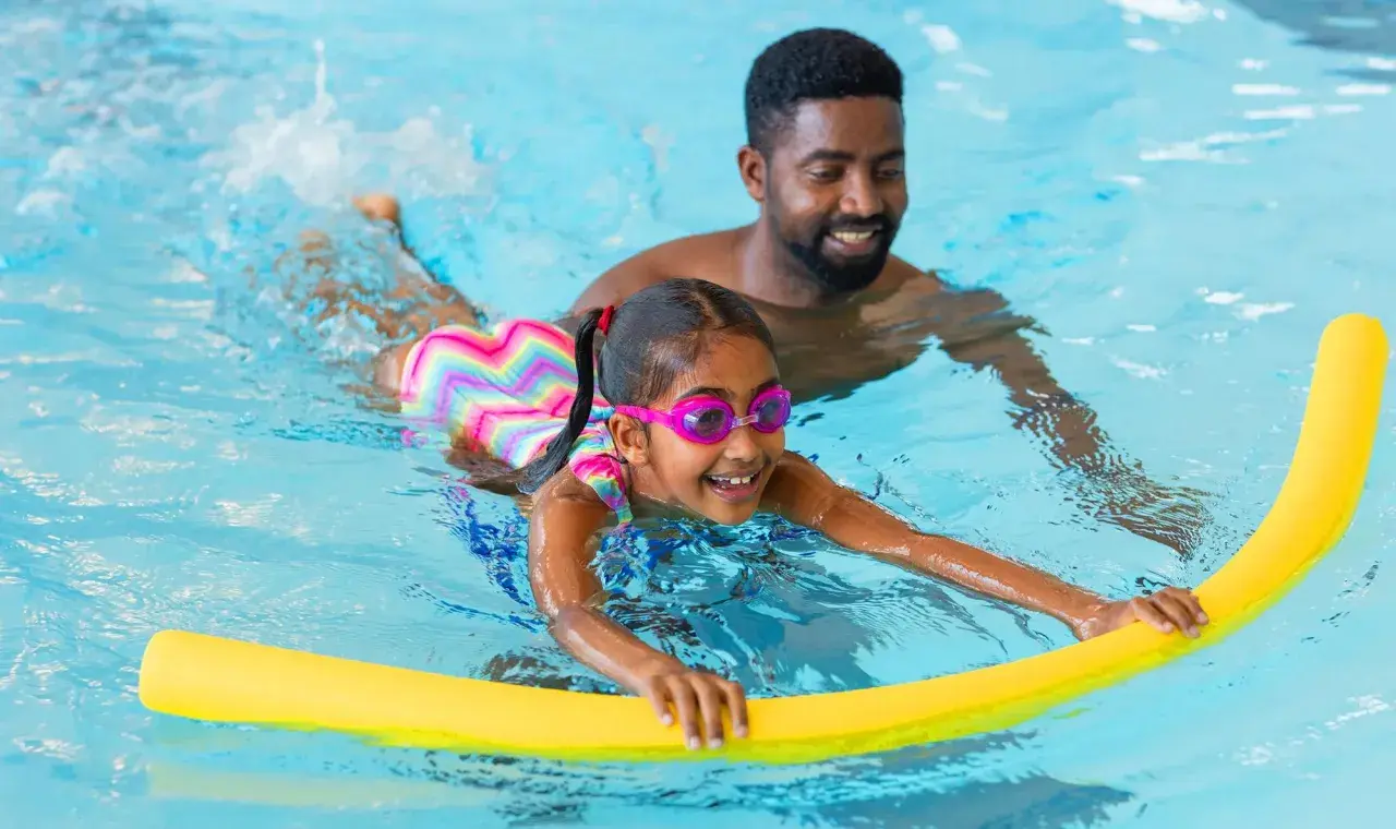 A father smiles as he helps his daughter with her swim lessons. She's wearing goggles and holding a yellow pool noodle.