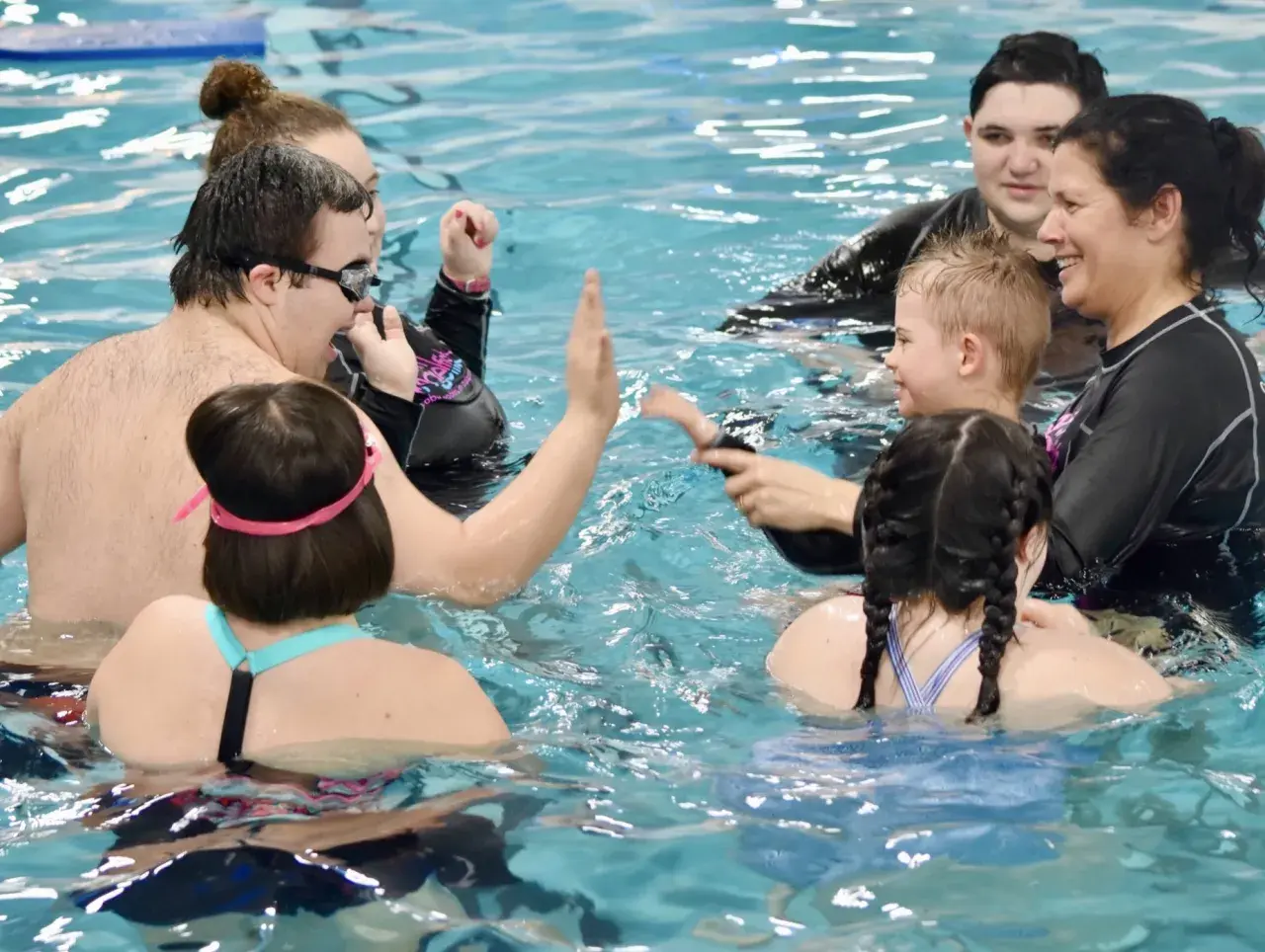 A group enjoys swim lessons for special needs, with instructors and participants high-fiving and smiling in the pool.