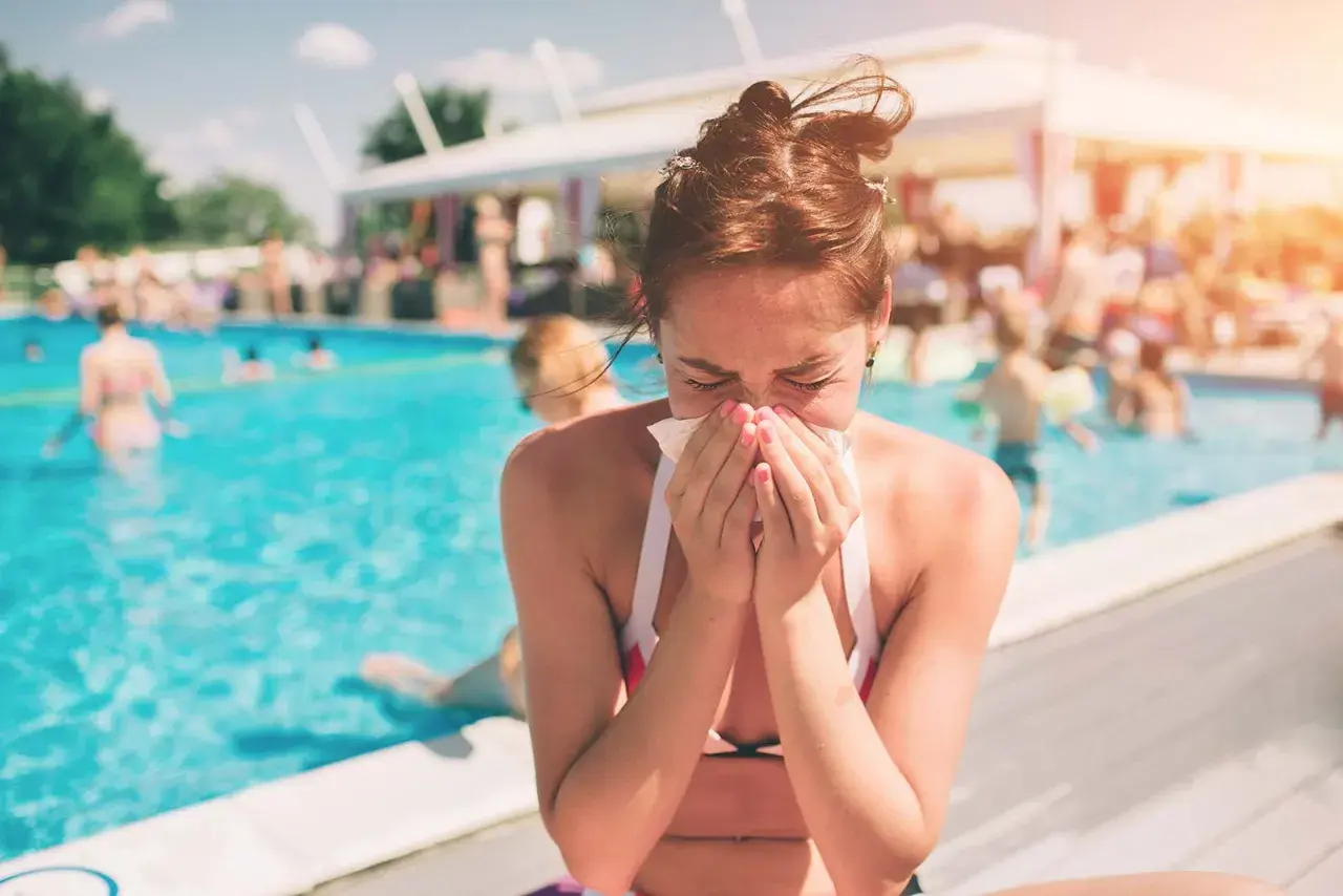 A woman with swimmer's sinusitis sneezes into a tissue by a crowded pool, unable to enjoy the summer day.