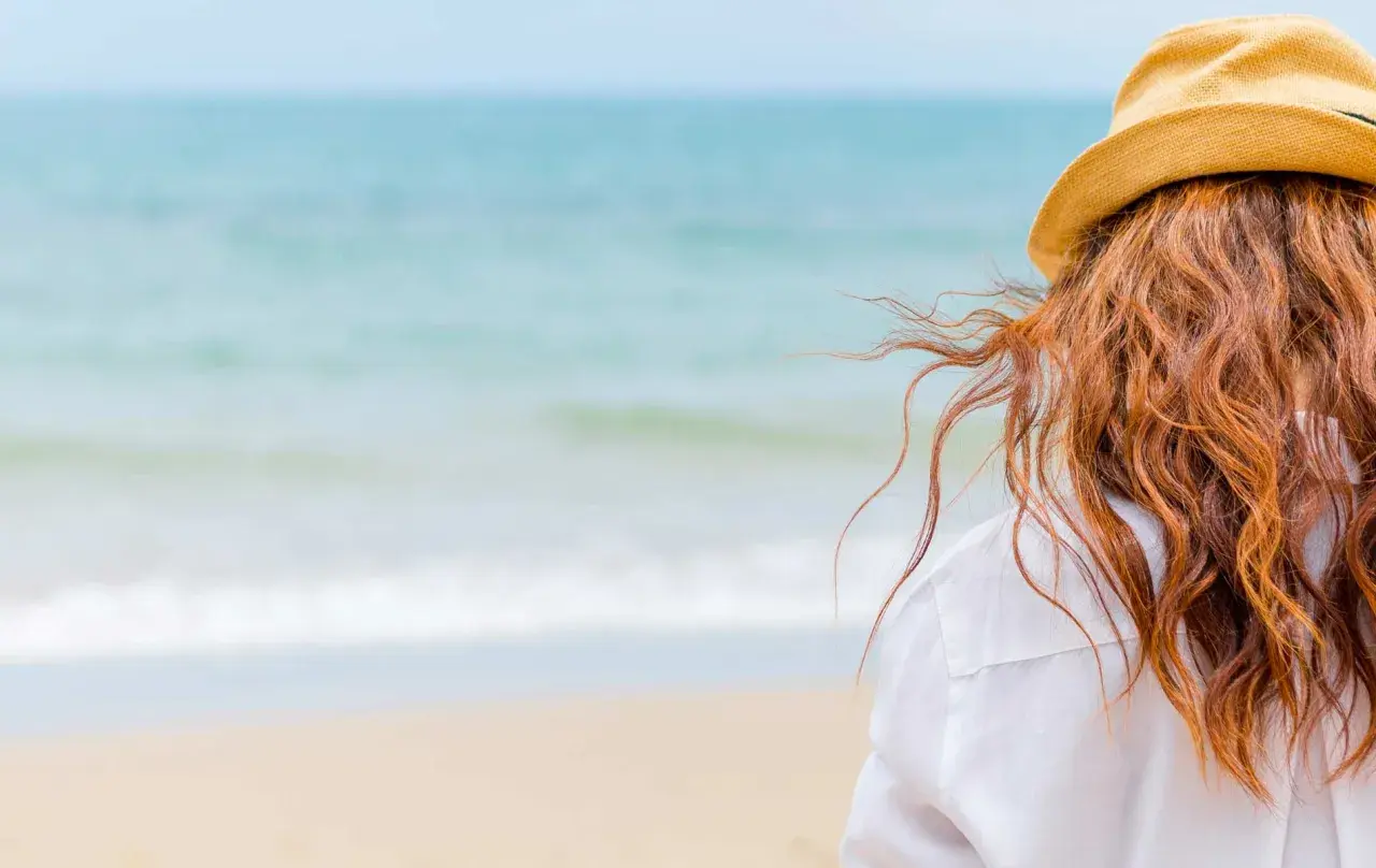 Woman with red hair in a straw hat on the beach, wondering how soon can I swim after coloring my hair.