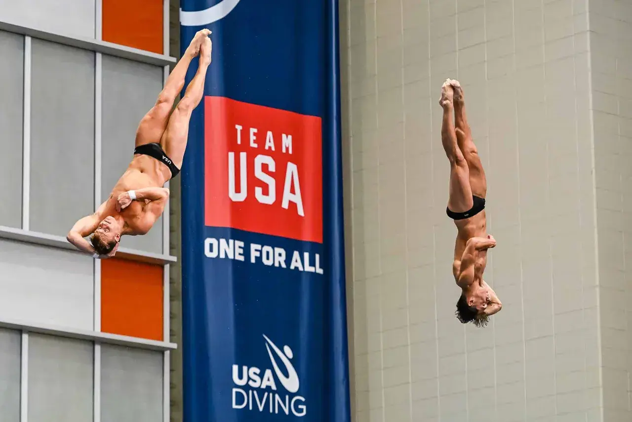 Two divers perform a synchronized high dive, suspended mid-air in a dramatic pose against a "Team USA" banner.