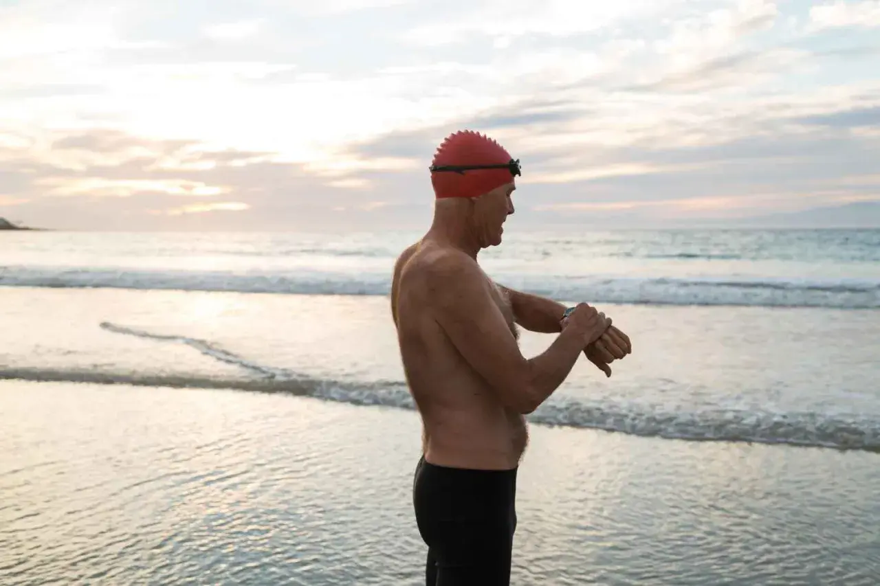 Man in swim cap checks watch on beach, ready for his swim. Early morning or late evening might be the best time to swim for weight loss.