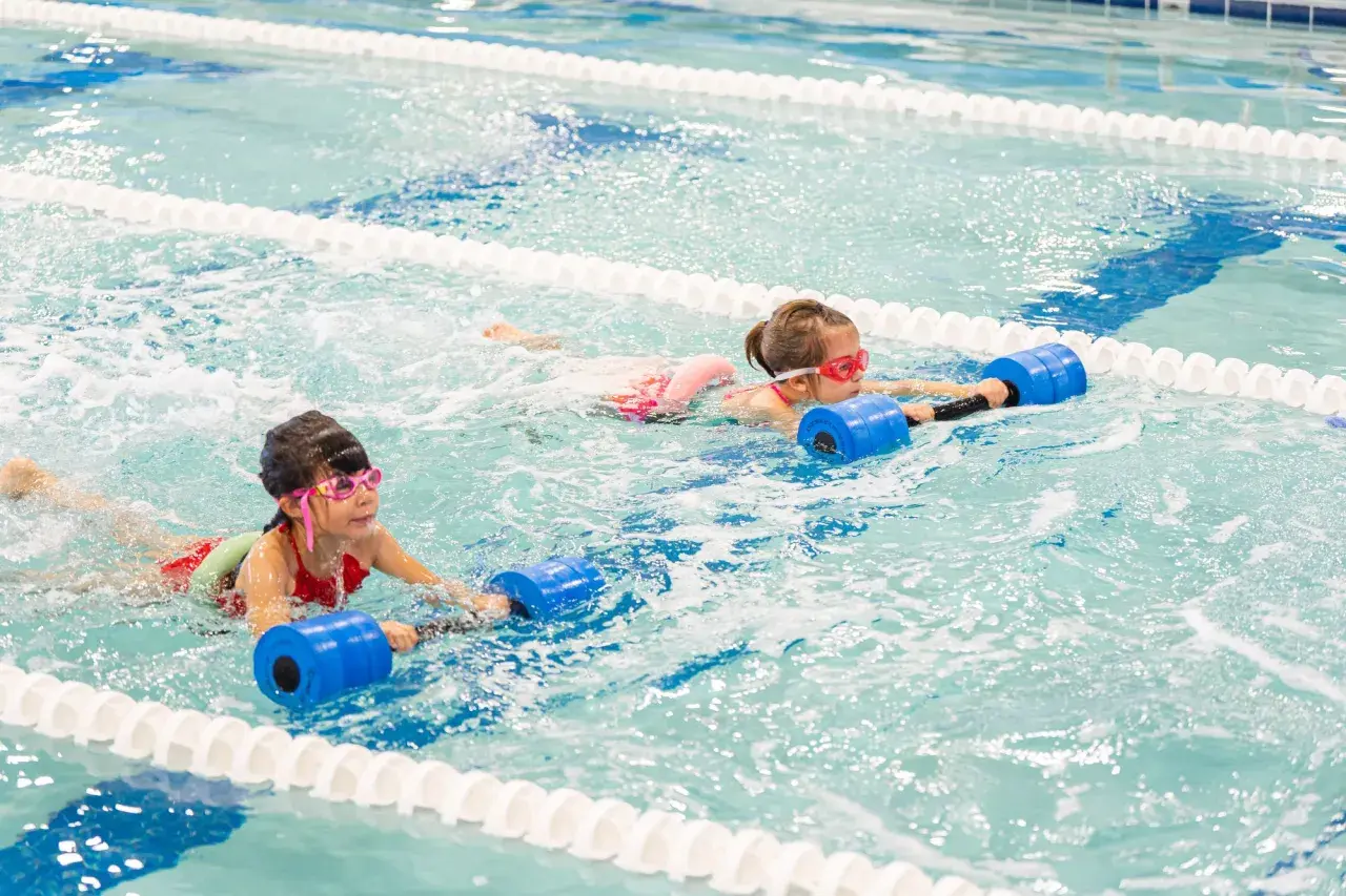 Two girls with goggles swim in a pool, holding blue flotation devices. One girl's face is partially submerged, showing her determination.