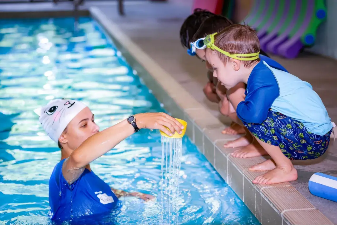 Instructor demonstrates water pouring during swim lessons for teens. Children watch intently from the poolside.