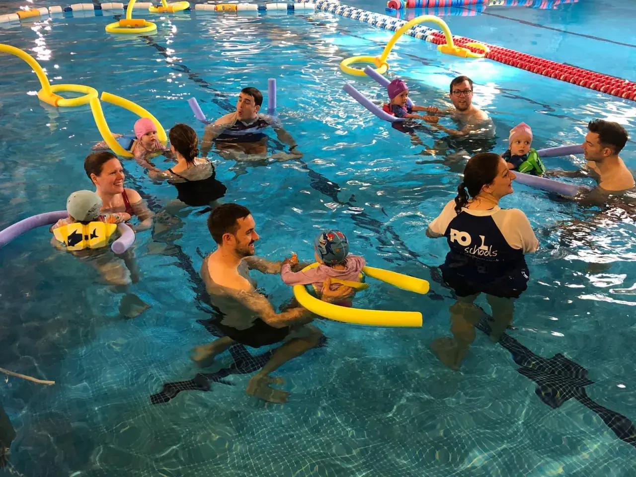 Parents and babies enjoy mommy and me swim classes, splashing and playing with pool noodles in the bright blue water.