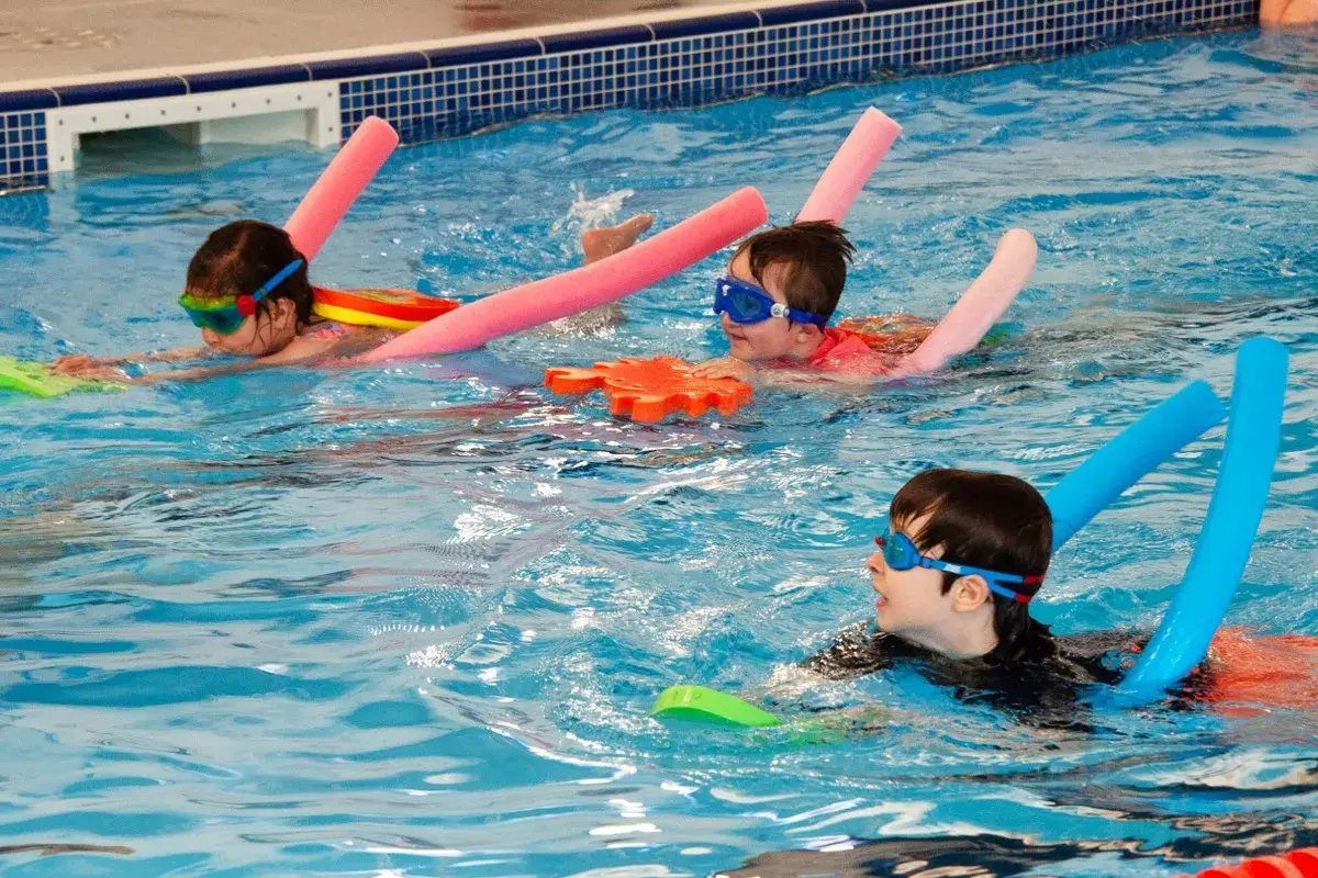 Children in goggles and swim aids enjoy a swim lesson, practicing their strokes in a bright blue pool.