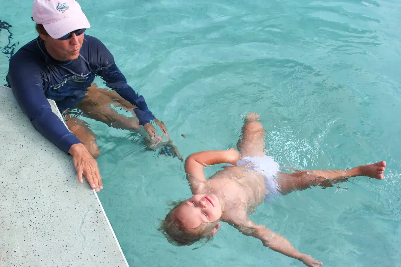 A child floats on their back in a pool, learning to swim. An instructor in a blue rash guard and pink hat watches.