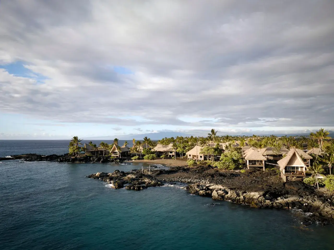 Tropical bungalows on a rocky coast in Kona, Hawaii. The calm ocean reflects the cloudy sky, a stark contrast to the power of a tsunami.