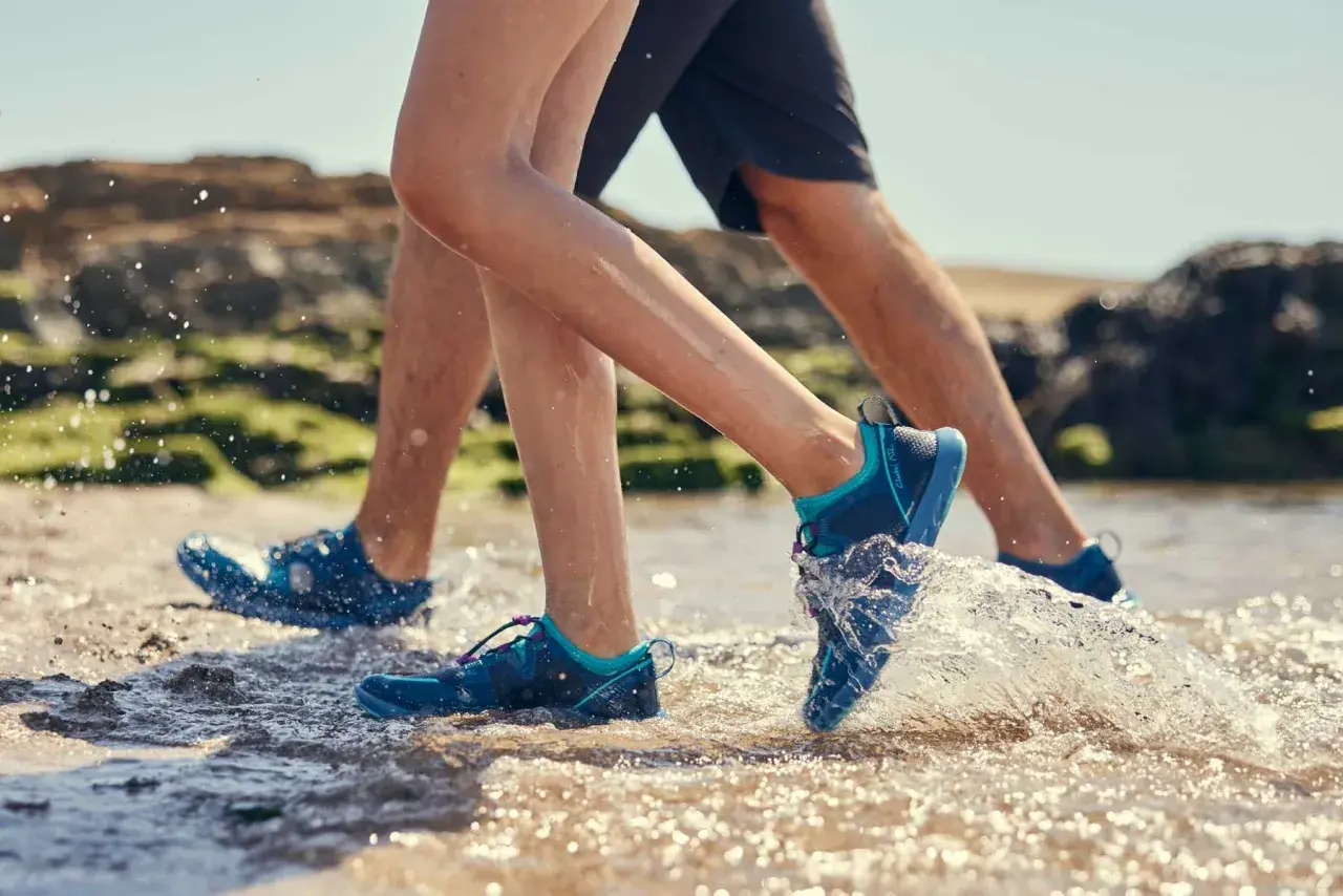 Two people wearing aqua jog shoes splash through shallow water on a rocky beach.