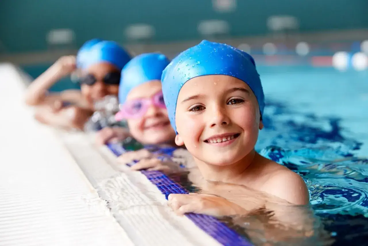 Happy kids in blue swim caps at swim lessons for kids, smiling by the pool edge.