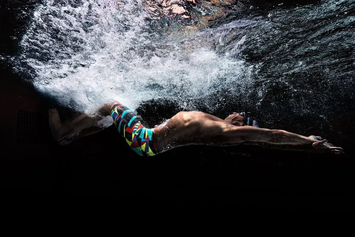 Underwater shot of a swimmer in a colorful tech suit, arms extended, with bubbles and light above.