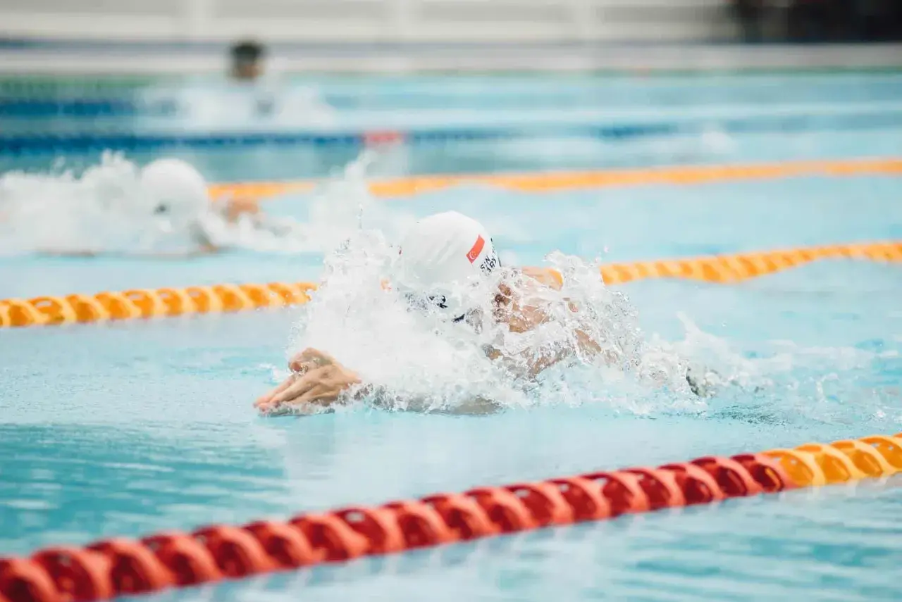 A swimmer in a white cap and goggles powers through the water, creating a splash. Their focused swimmer posture is evident as they propel forward in the lane.