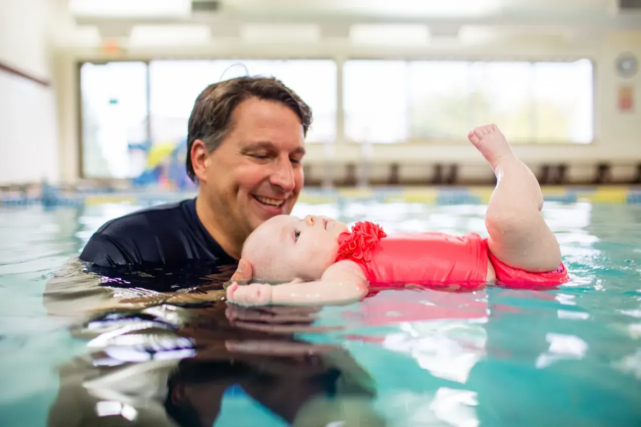 A father smiles as he holds his baby in a pool, showing that kids can learn to swim from a very young age.