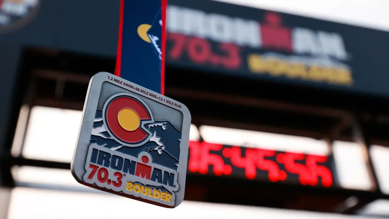 Ironman 70.3 Boulder medal hangs in front of a finish line clock. The medal features the Colorado flag and mountains.