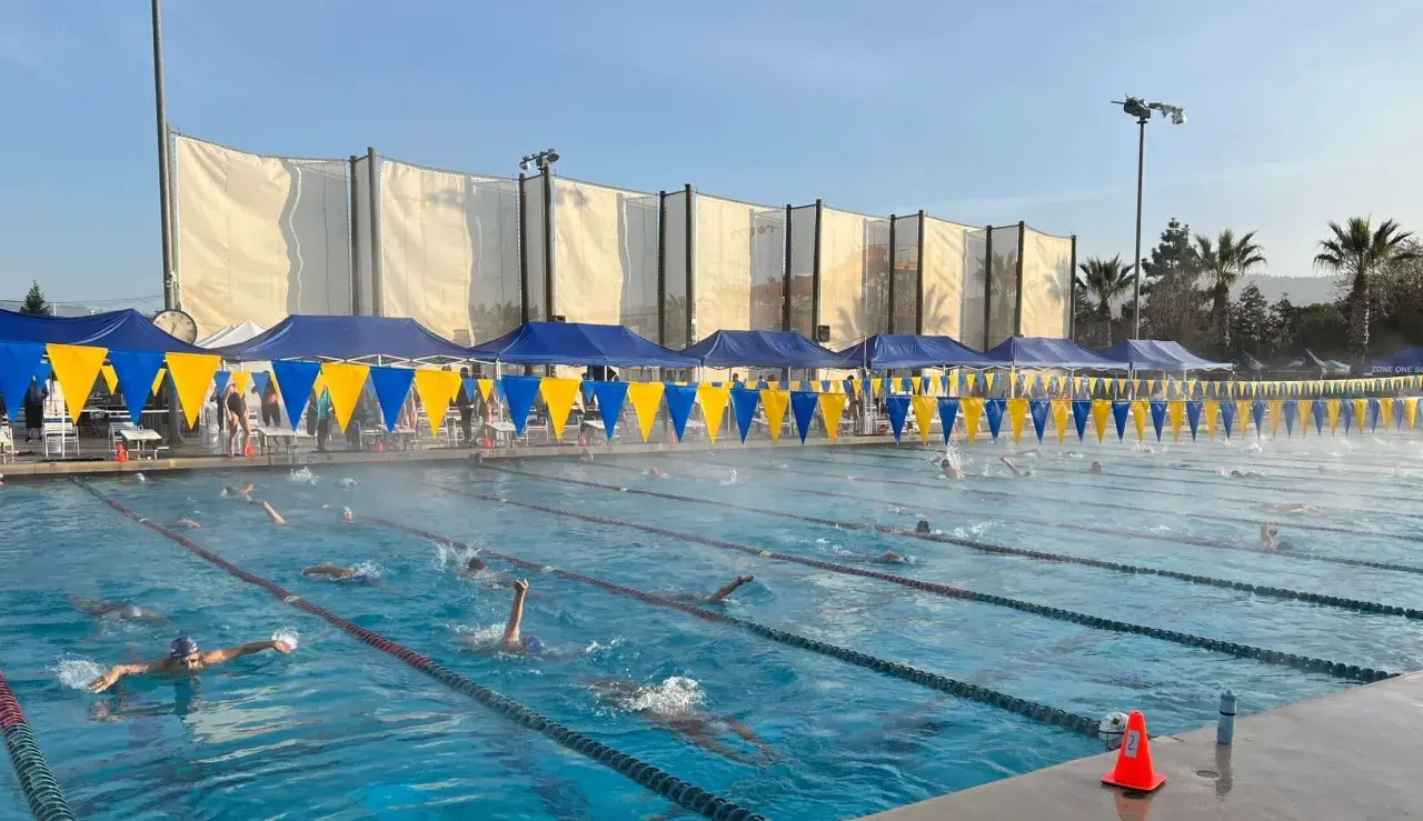 Swimmers race in lanes during a circle swim event. Blue and yellow banners line the pool, with tents and spectators in the background.
