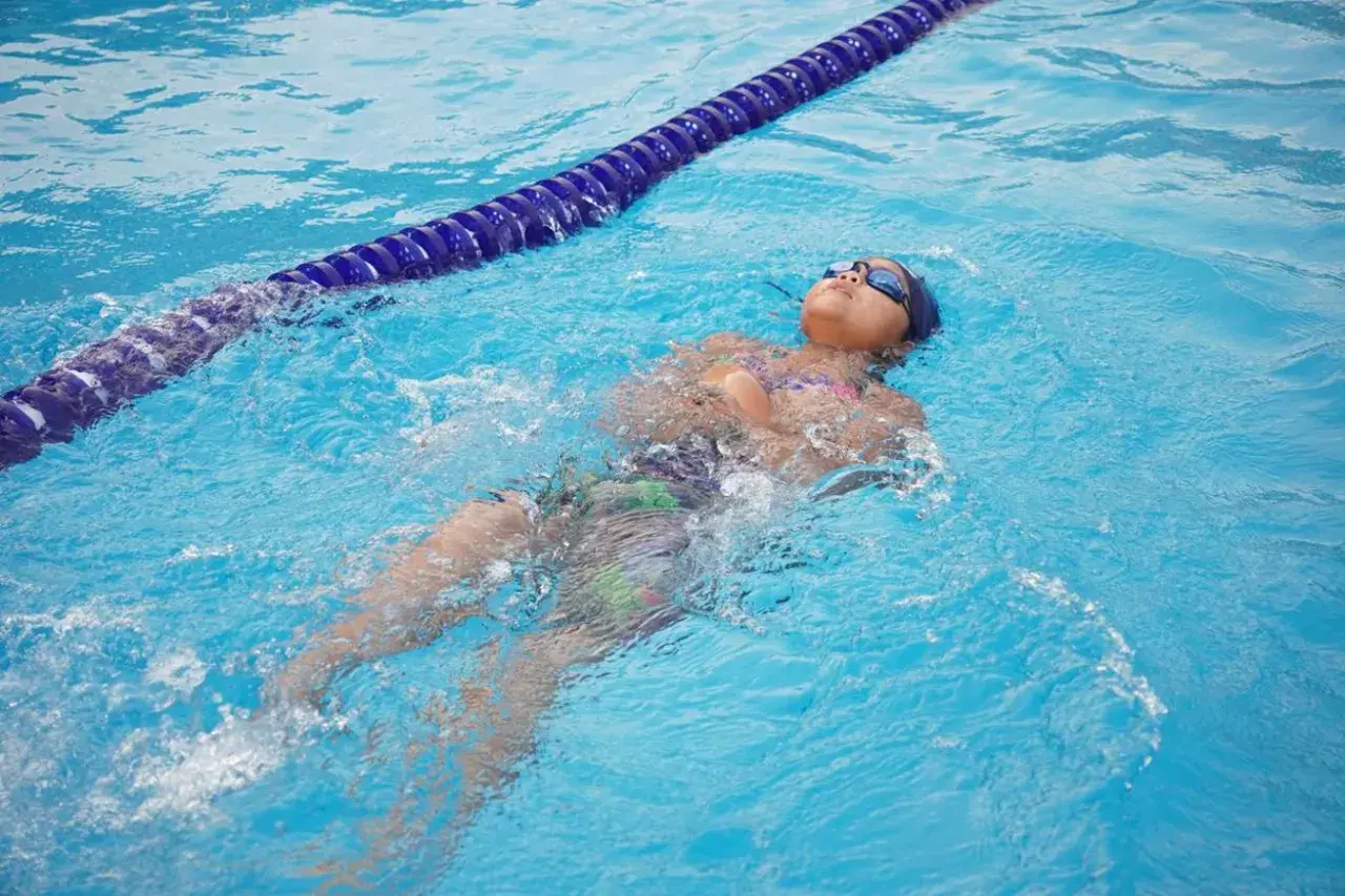 A child practices how to tread water in a pool, floating on their back with goggles on.