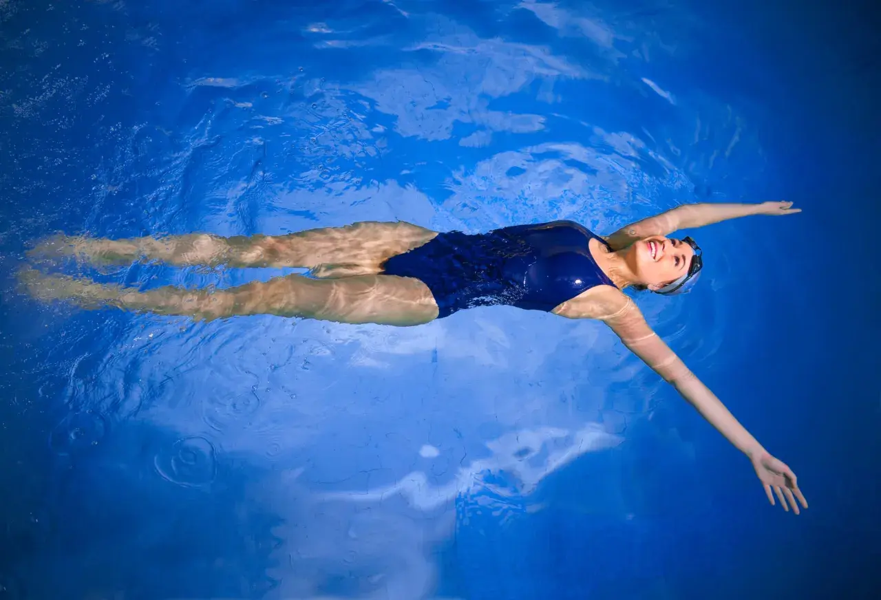 A woman in a blue swimsuit floats on her back in a pool, smiling. It makes you wonder, can everyone float like this?
