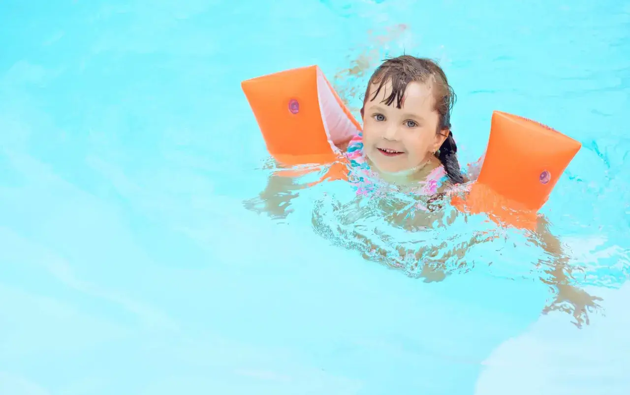A happy young girl with bright blue eyes is floating in the pool, wearing orange arm floaties and a colorful swimsuit.
