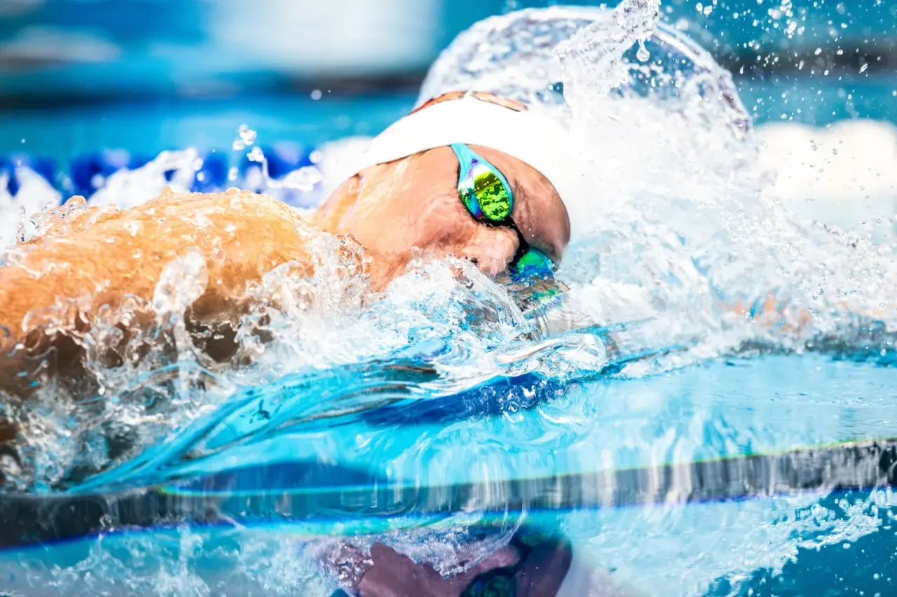 A swimmer's head emerges from the water, goggles reflecting the blue pool. Water splashes around them, a dynamic scene of fun swim drills.