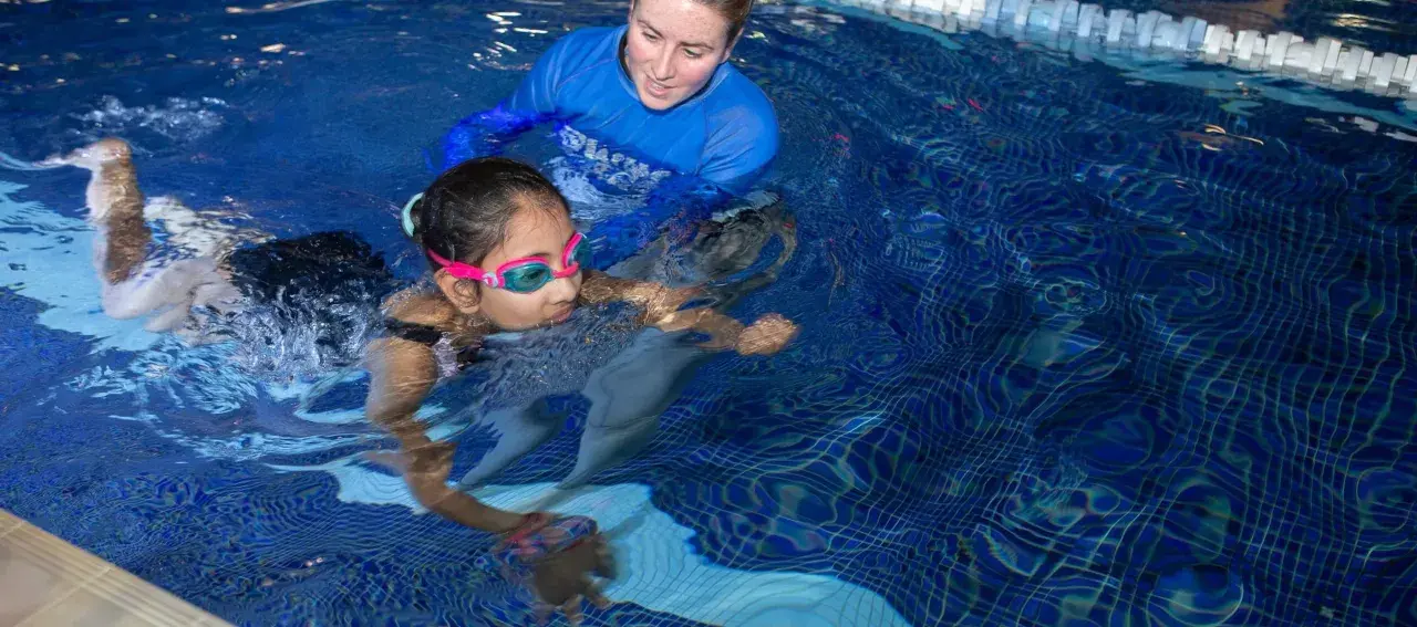 A swim instructor guides a young girl learning to swim. The question of how long does it take to learn how to swim varies, but with practice, she'll be swimming confidently.