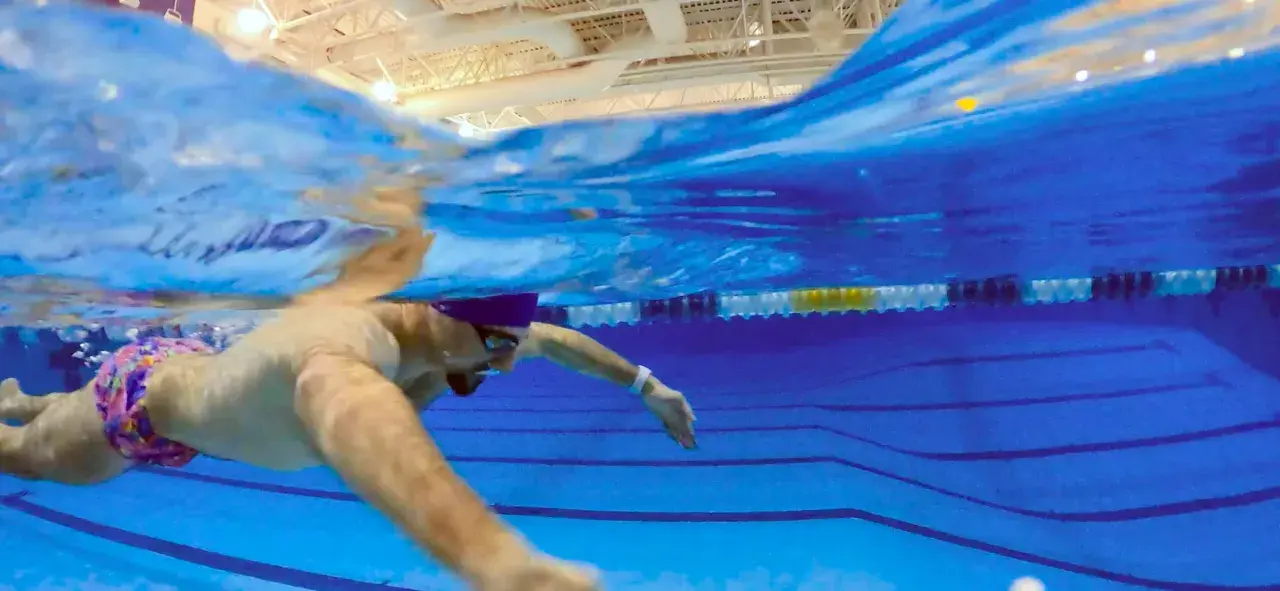 Underwater view of a swimmer in colorful trunks, demonstrating what is sculling with their arms to propel themselves through the blue pool water.