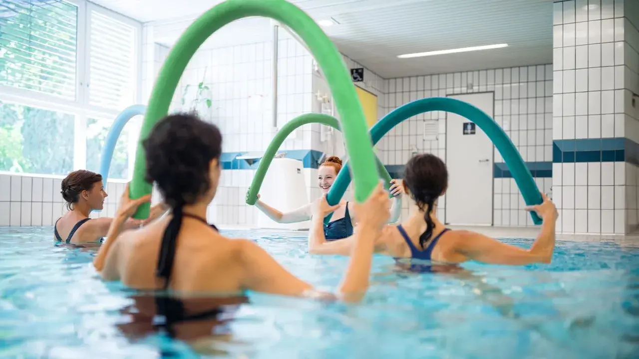 Women enjoy an aqua zumba class, holding colorful pool noodles for resistance and fun in the water.
