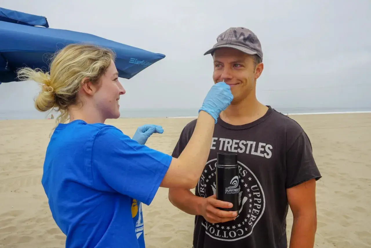 A woman in a blue shirt and gloves gently swabs a man's nose on a beach. He's wearing a cap and a t-shirt that reads "Trestles." This is part of a study to see if you can swim with a staph infection.