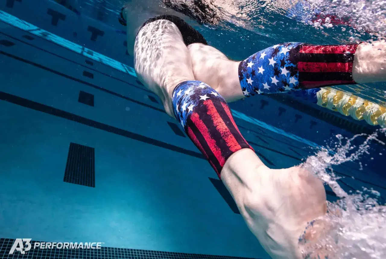 Underwater view of a swimmer's legs in a pool, wearing patriotic-themed compression sleeves. Wondering why do I get foot cramps in the pool?