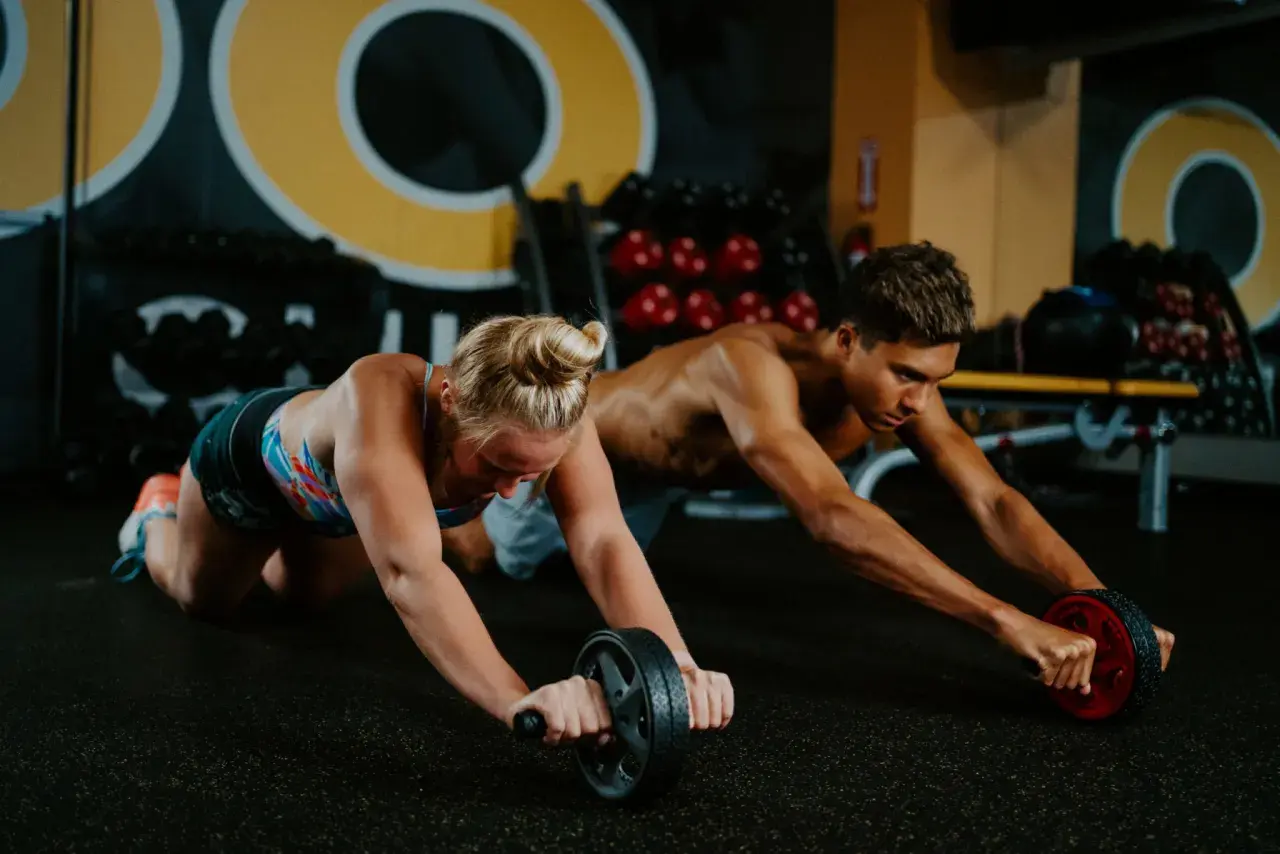 Two athletes, like swimmers training, perform ab roller exercises in a gym.
