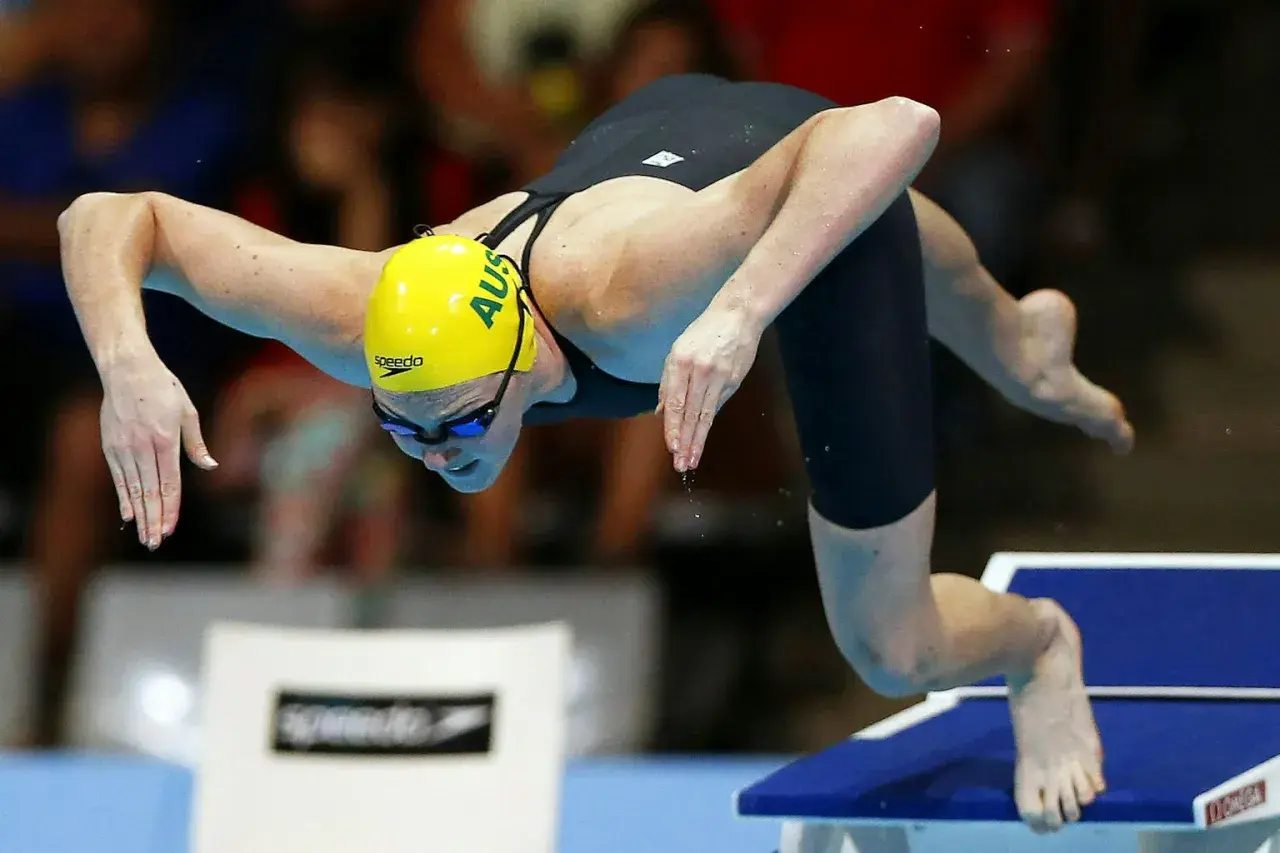 An Australian swimmer in a yellow cap prepares for a powerful high diving start, poised on the edge of the block.