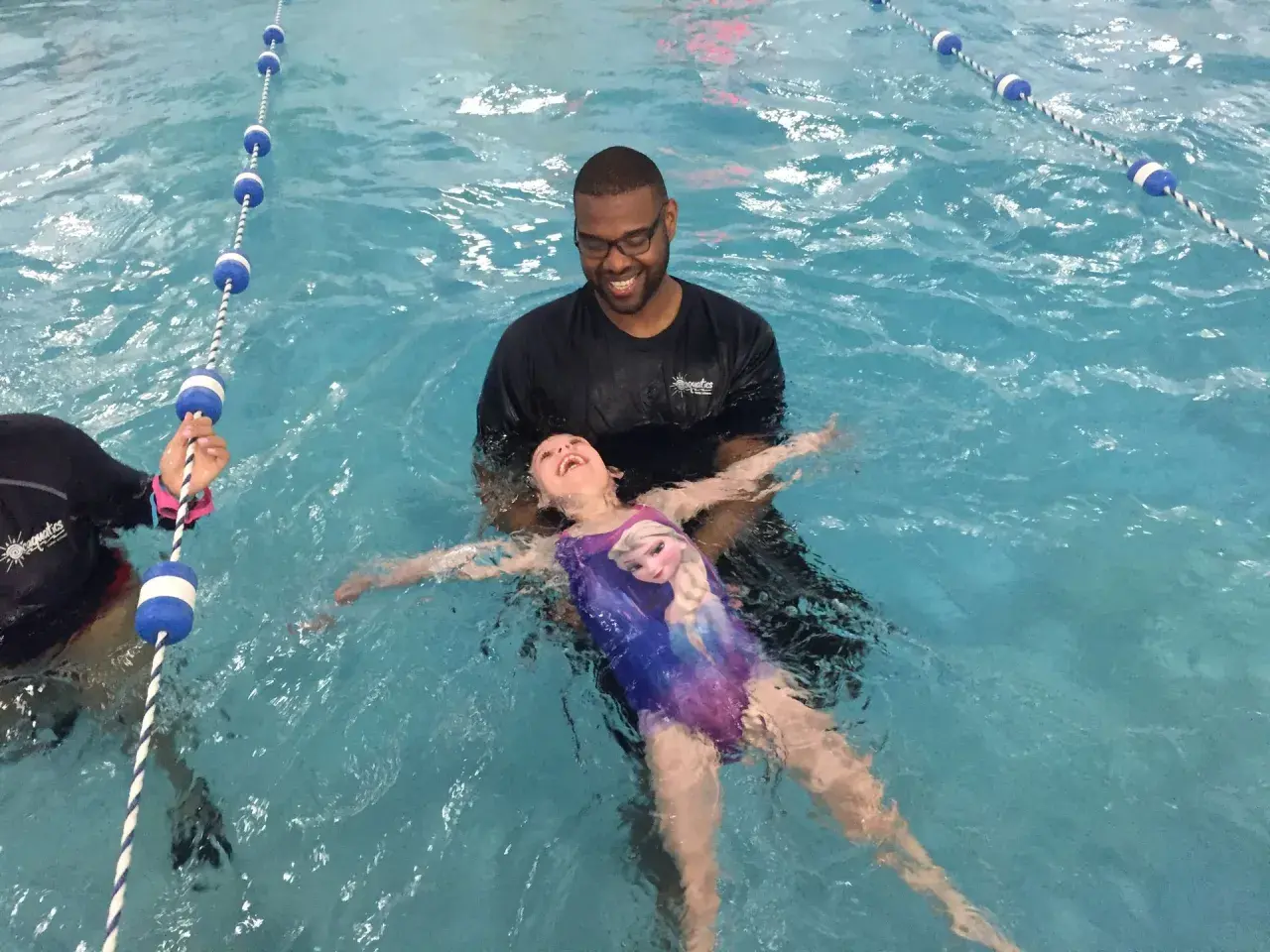 A smiling instructor helps a young girl in a Frozen swimsuit learn how to float on her back in a swimming pool.