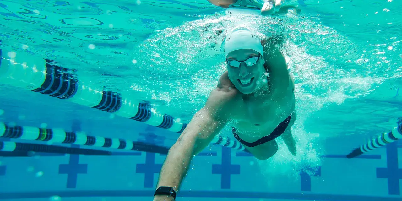 A smiling man swims underwater in a pool, enjoying easy swim workouts. Lane lines and pool markings are visible.