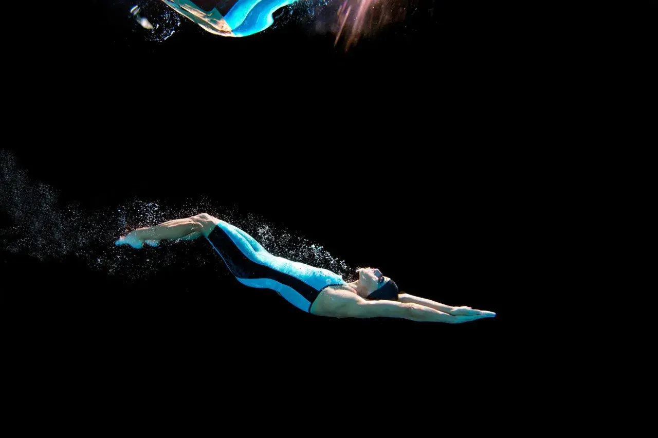 Swimmer in a blue and black suit performs a powerful dolphin kick underwater, creating a trail of bubbles against a dark background.