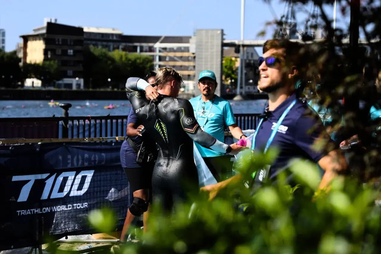 A triathlete in a wetsuit is helped by staff during a triathlong transition. The T100 Triathlon World Tour banner is visible.