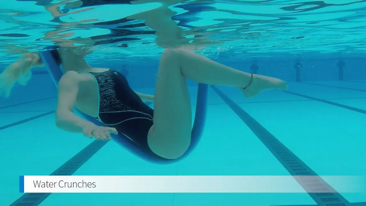 Woman performing water crunches in a pool, using a pool noodle for support. The water's surface reflects the light above.