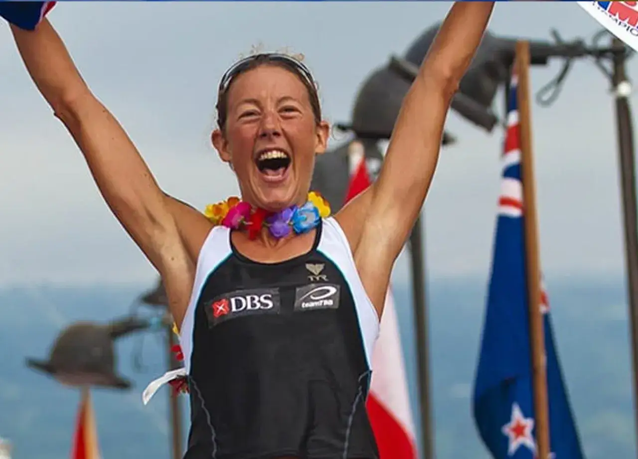 Chrissie Wellington celebrates a victory, arms raised, wearing a colorful lei and a racing singlet.