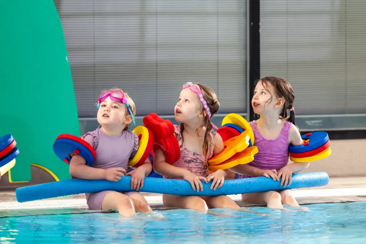 Three young girls in goggles and swimsuits sit at the edge of a pool, holding colorful flotation devices, ready for their swim lessons.