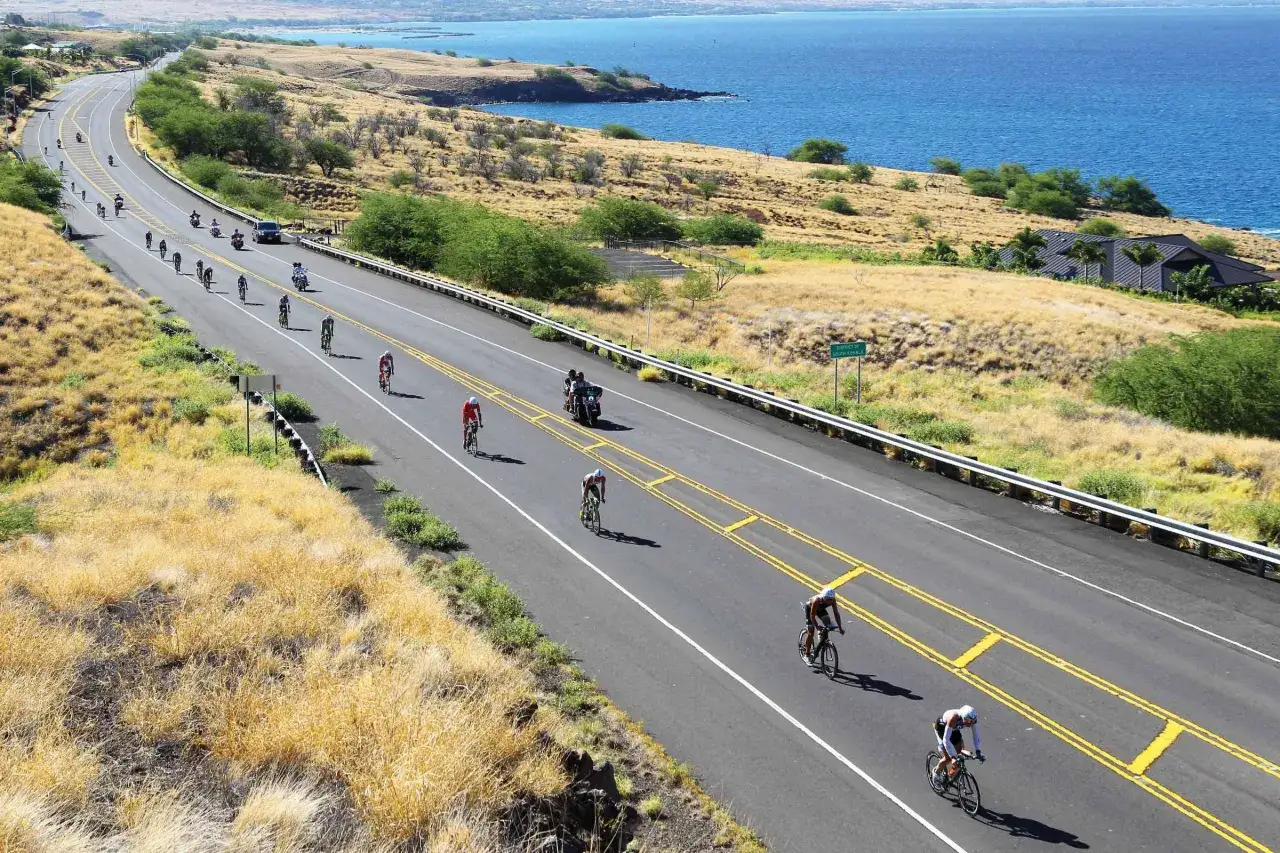Cyclists race along a coastal road in Hawaii during the Hawaii 70.3 triathlon, with the blue ocean in the background.