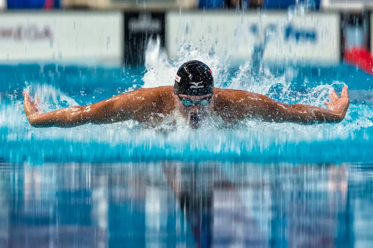 A swimmer in a black cap and goggles performs a powerful butterfly stroke, mid-breath, creating a dramatic splash.
