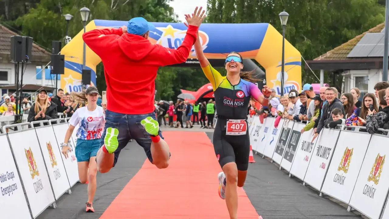 A triumphant woman crosses the finish line of an Ironman race, her colorful tri-suit a blur of motion. A man in a red hoodie jumps for joy beside her, celebrating her incredible achievement.