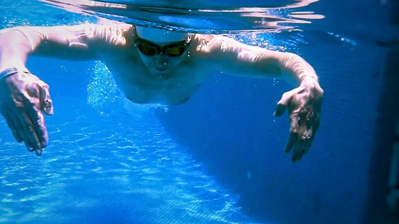 Underwater view of a swimmer wearing goggles, demonstrating the sculling technique with hands moving to propel through the blue water.