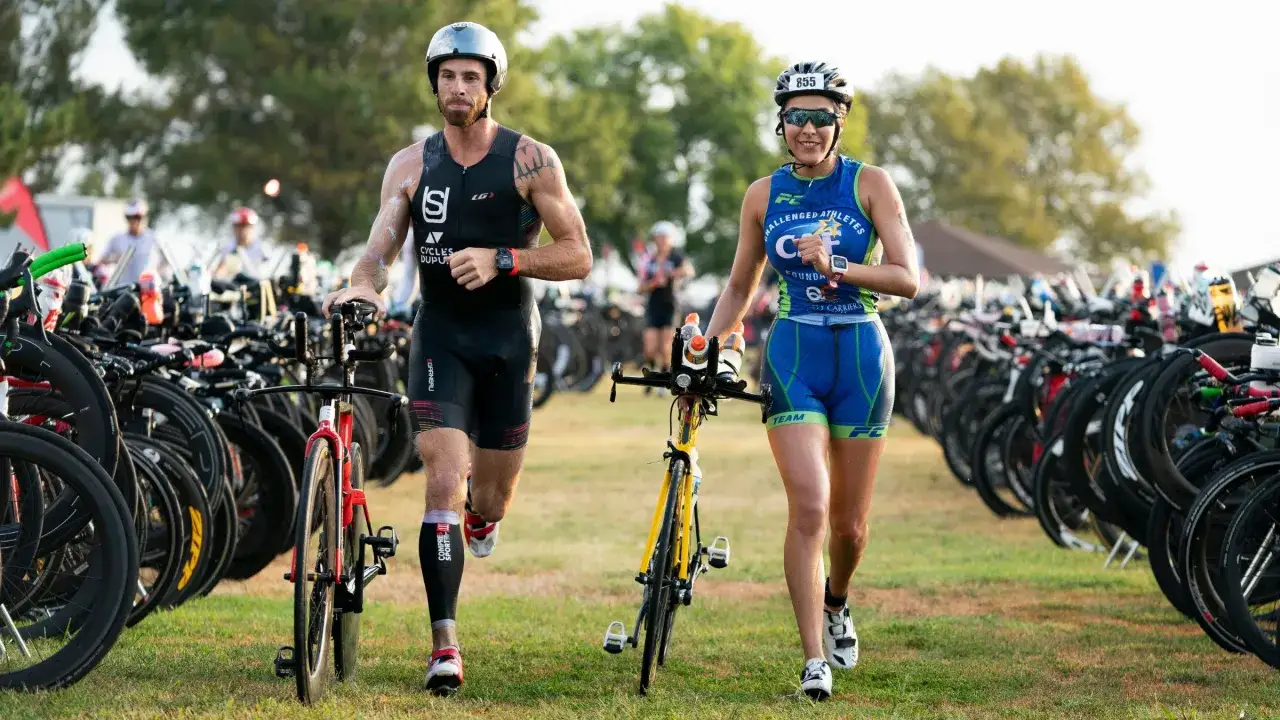 A male and female athlete, ready for their team triathlon, jog past rows of bicycles.
