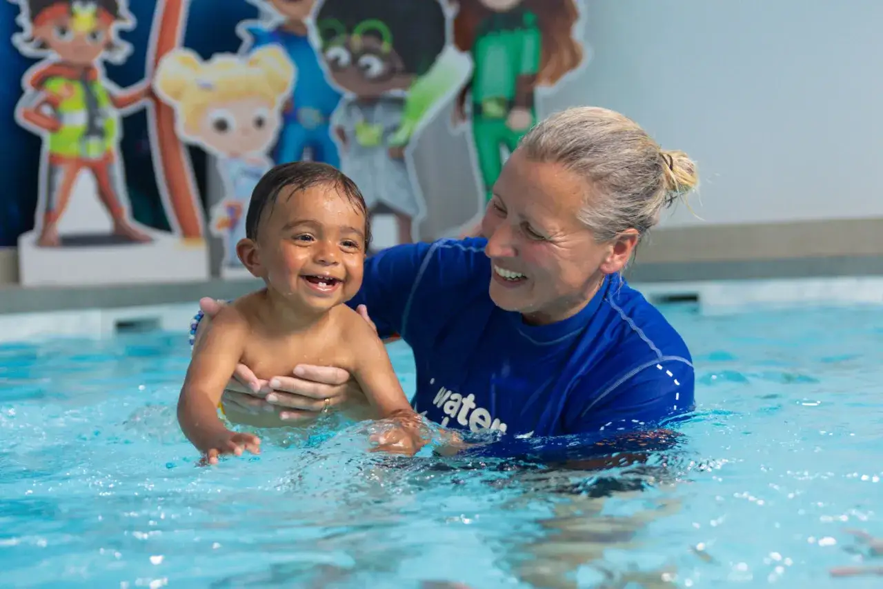 A smiling toddler enjoys a swim lesson, held by an instructor in a blue shirt.