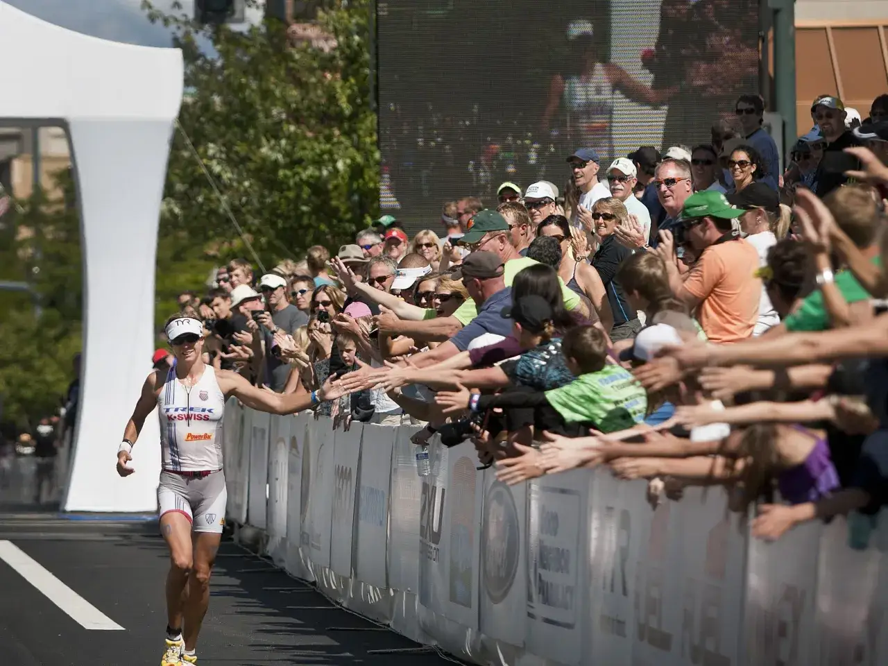 A female athlete runs towards the Ironman finish line, reaching out to high-five cheering spectators.