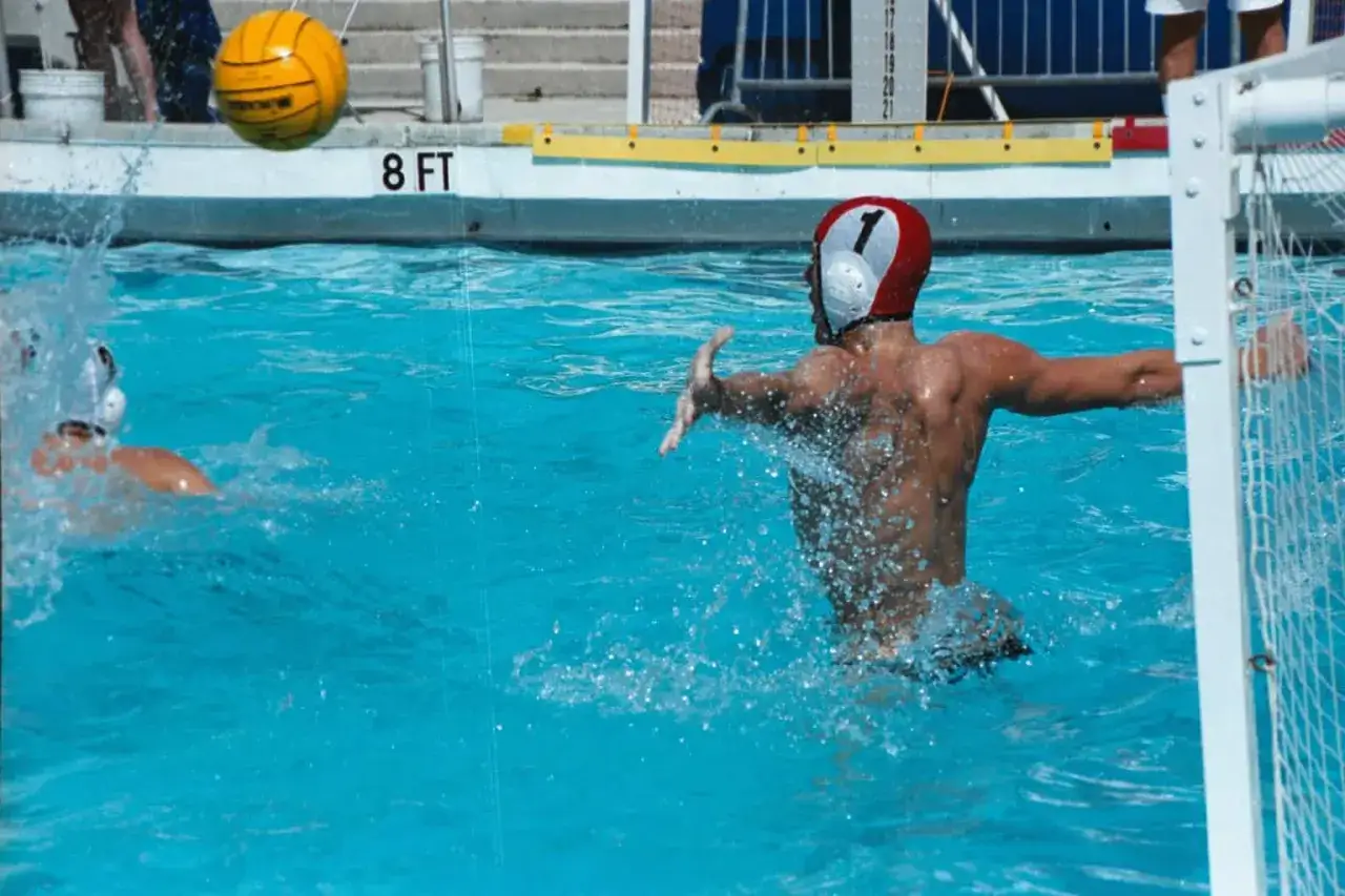 Water polo players treading water, one with a red and white cap numbered 1, splashes as they defend the goal.