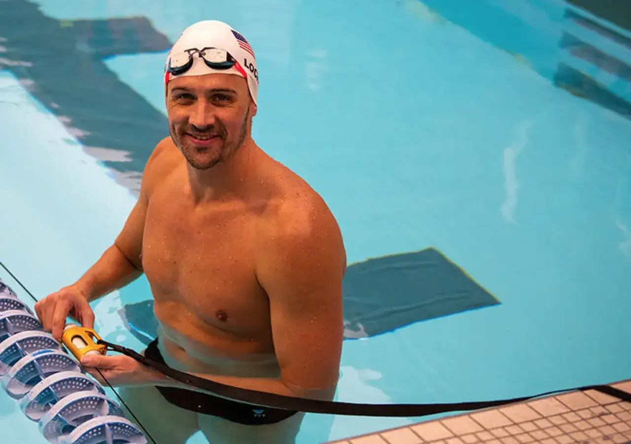 Swimmer Ryan Lochte, wearing a cap and goggles, smiles in a pool, ready for his intense Ryan Lochte workouts.