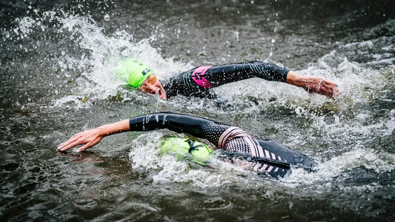 Two swimmers in Ironman wetsuits race through choppy water, their neon green caps bobbing. Following Ironman wetsuit rules, they push forward.