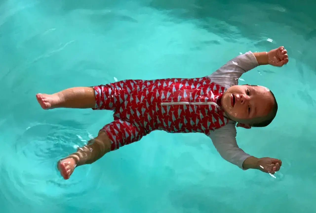 A baby in a red swimsuit with fish patterns floats on their back during ISR swim lessons.